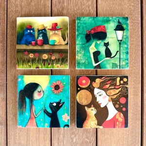 以下が含まれることがあります： 猫と人間のイラストが描かれた、正方形のアートプリント4枚。1枚は帽子をかぶりマグカップを持っている3匹の猫、もう1枚は黒猫と女性、残りの2枚は花を持つ少女と猫と女性が描かれています。
