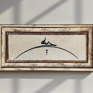 May include: Rectangular framed artwork with a textured off-white background. The frame is distressed with a light brown and cream color. Black calligraphy is centered on the artwork, with a curved line and dots.