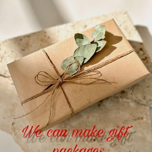 May include: A wrapped gift box in brown paper, tied with twine and adorned with a sprig of green leaves. The text "We can make gift packages" is written in red cursive on the surface.