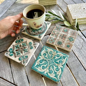 May include: A collection of square ceramic coasters with intricate, embossed designs in teal, brown, and white. One coaster holds a coffee-filled mug. The coasters are arranged on a weathered wooden surface, suggesting a rustic or Mediterranean theme.