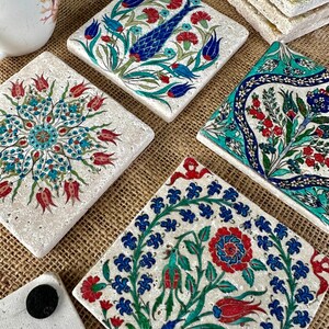 May include: A collection of square stone coasters with intricate floral designs in red, blue, and green. The coasters feature various patterns, including stylized flowers and a fish motif, set against a neutral background. A cup of coffee is visible in the background.