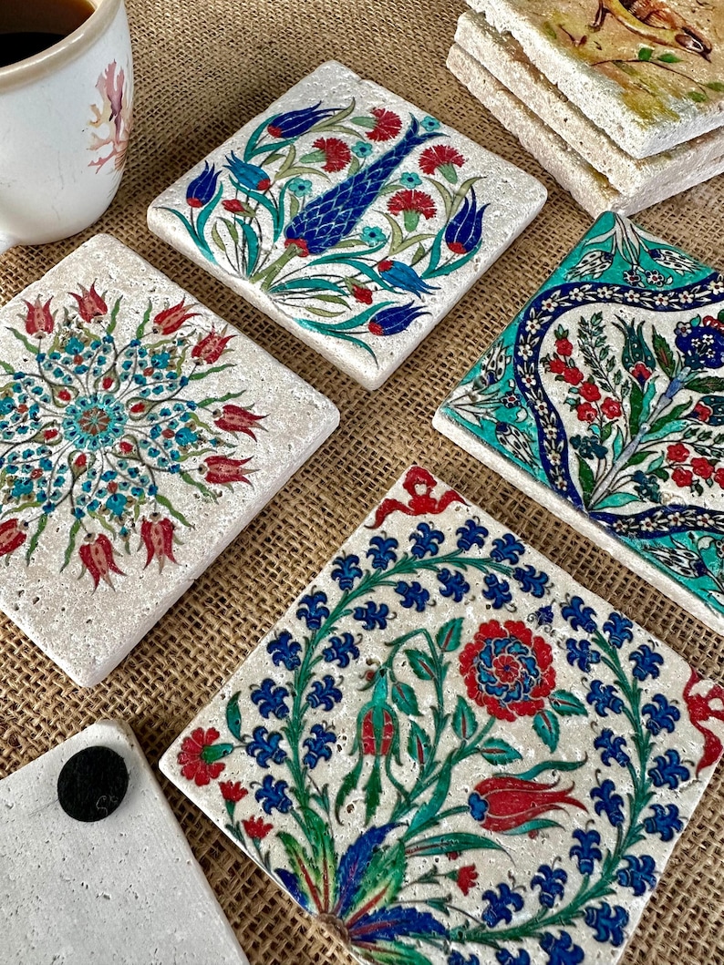 May include: A collection of square stone coasters with intricate floral designs in red, blue, and green. The coasters feature various patterns, including stylized flowers and a fish motif, set against a neutral background. A cup of coffee is visible in the background.