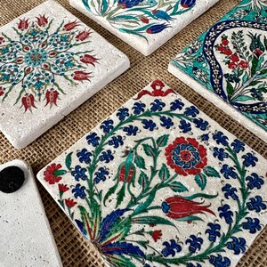 May include: A collection of square, decorative coasters with intricate floral designs in red, blue, and green on a white background. One coaster has a white mug with a floral design on top. The coasters are made of a textured material.