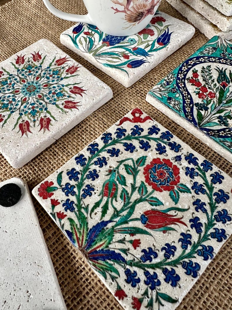 May include: A collection of square, decorative coasters with intricate floral designs in red, blue, and green on a white background. One coaster has a white mug with a floral design on top. The coasters are made of a textured material.