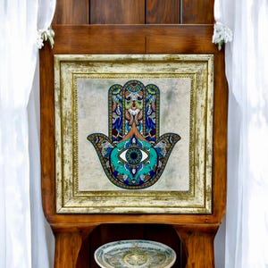 May include: A framed artwork featuring a colorful Hamsa hand with a blue eye in the center. The Hamsa is a popular symbol of protection and good luck in many cultures.