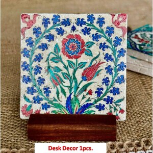 May include: Square desk decor with a floral design in red, blue, and green on a cream background. The design features a central flower and surrounding leaves and blooms. It is displayed on a dark brown wooden stand.