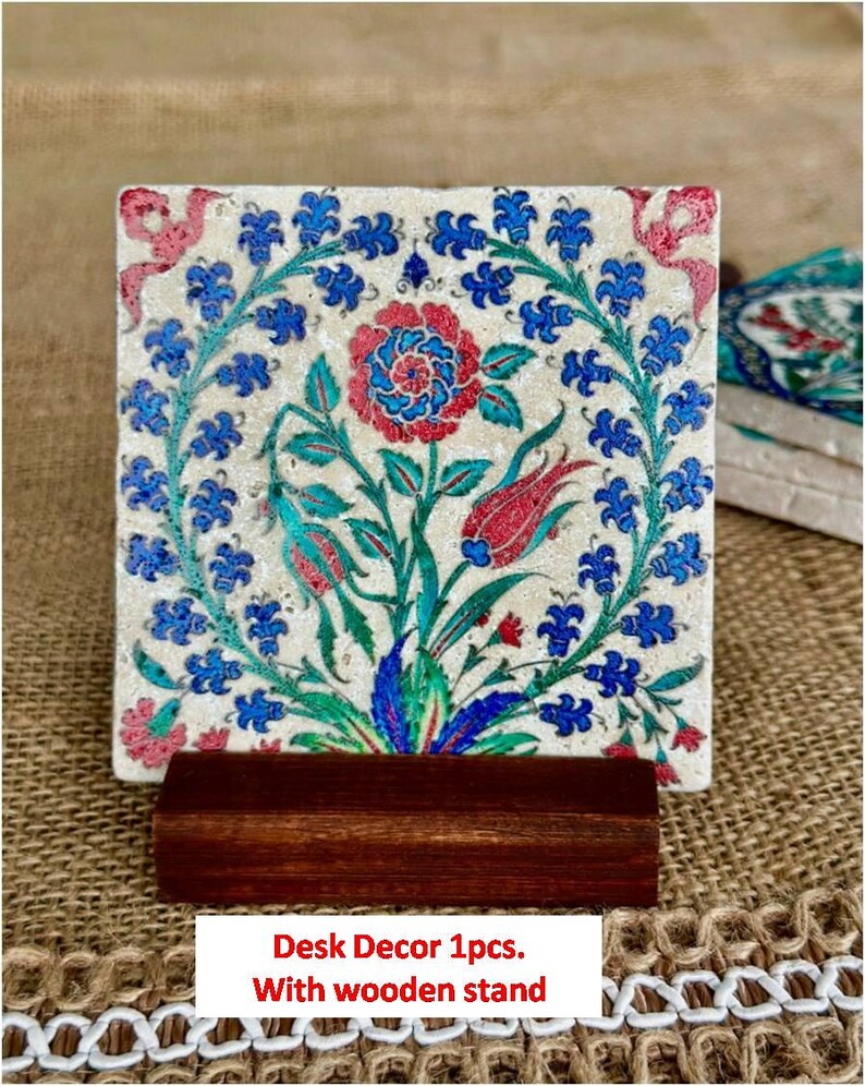 May include: Square desk decor with a floral design in red, blue, and green on a cream background. The design features a central flower and surrounding leaves and blooms. It is displayed on a dark brown wooden stand.