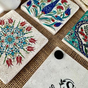May include: Square stone coasters with colorful floral designs in red, blue, and turquoise. The coasters have a textured, off-white surface. One coaster has the text "STONEA by Levi" with a logo. The coasters are on a brown surface.