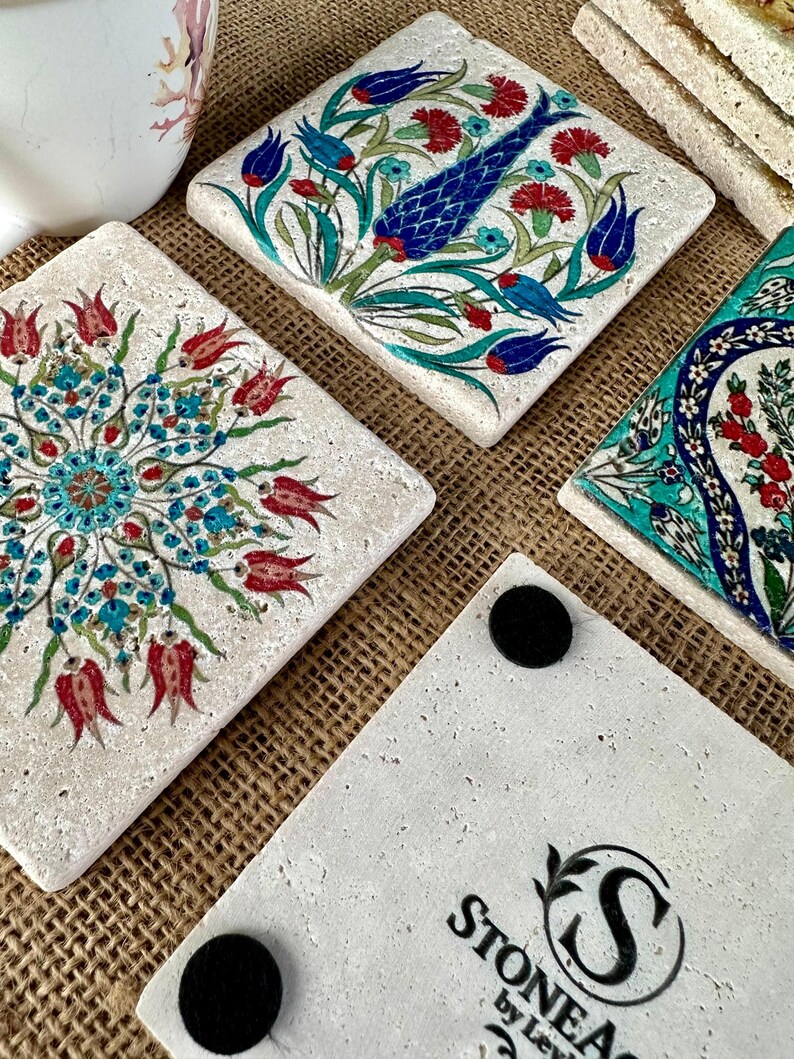 May include: Square stone coasters with colorful floral designs in red, blue, and turquoise. The coasters have a textured, off-white surface. One coaster has the text "STONEA by Levi" with a logo. The coasters are on a brown surface.