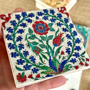 May include: Square ceramic coaster with a floral design. The coaster features a central red and blue flower surrounded by blue floral accents and green leaves. Additional coasters are visible in the background.