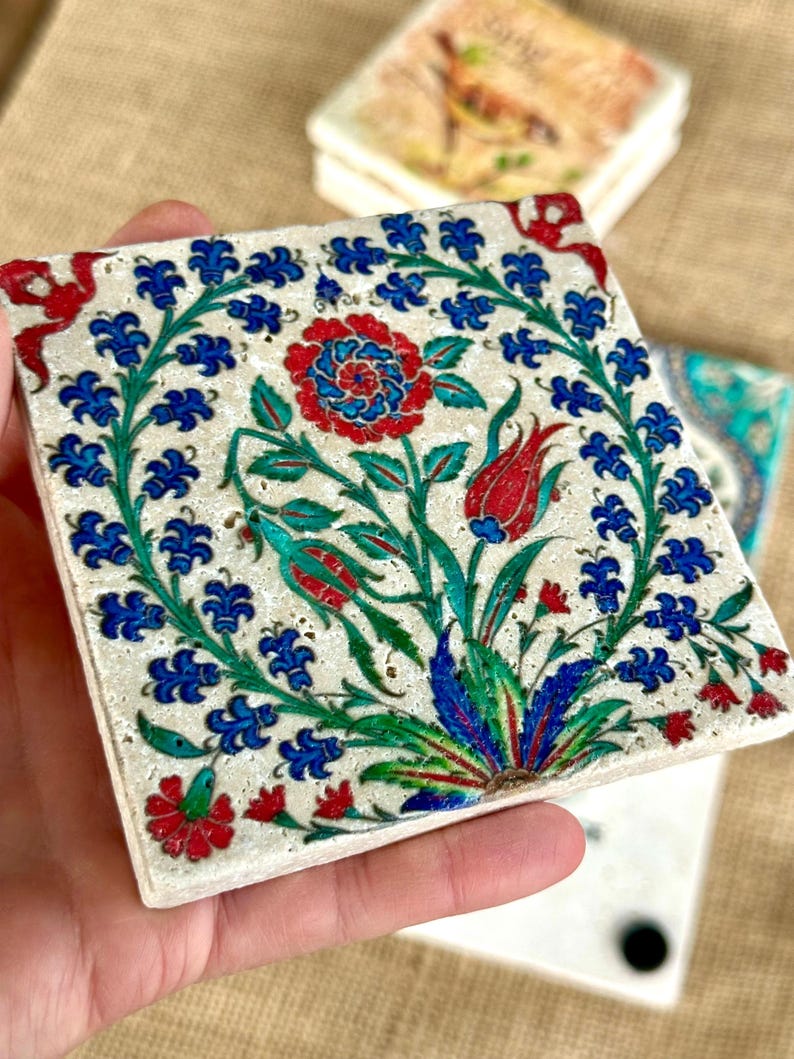 May include: Square ceramic coaster with a floral design. The coaster features a central red and blue flower surrounded by blue floral accents and green leaves. Additional coasters are visible in the background.