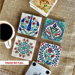 May include: A set of four square coasters with colorful floral designs. Each coaster features a unique pattern in shades of blue, red, and green. The coasters are arranged on a textured, tan surface, with a cup of coffee visible.