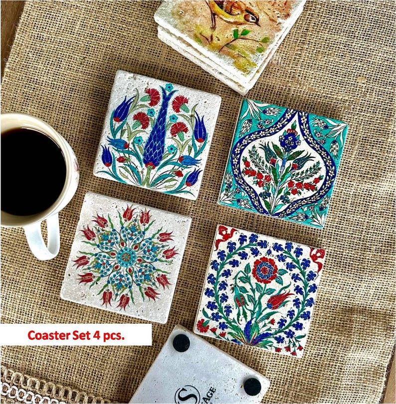May include: A set of four square coasters with colorful floral designs. Each coaster features a unique pattern in shades of blue, red, and green. The coasters are arranged on a textured, tan surface, with a cup of coffee visible.
