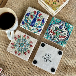 May include: Set of square stone coasters with colorful floral designs. The coasters feature red, blue, and turquoise patterns. One coaster has the text "STONEAGE by Leyla". A white mug with coffee is in the image.