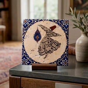 May include: A square decorative tile featuring a beige travertine-like center with black Arabic calligraphy in the shape of a whirling dervish. The border is decorated with a blue and white floral pattern, and the tile rests on a wooden stand.