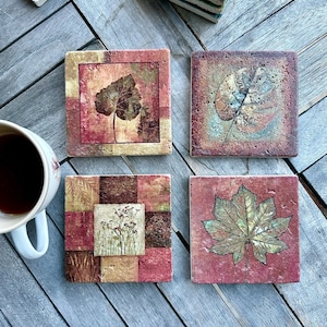Könnte beinhalten: Vier quadratische Untersetzer mit Blatt- und Blumendesigns. Die Untersetzer weisen eine Mischung aus Farben auf, darunter Rot, Braun und Gold. Die Untersetzer bestehen aus einem strukturierten Material und sind auf einer Holzoberfläche angeordnet.