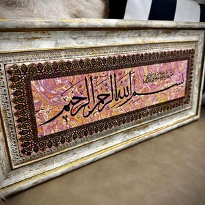 Op de afbeelding: Een ingelijst kalligrafisch kunstwerk met een roze en gouden marmeren achtergrond. Het kunstwerk toont Arabisch schrift in zwarte inkt, met de tekst "سبحان الله والحمد لله ولا اله الا الله والله اكبر".