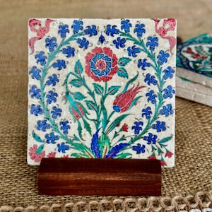 May include: Square ceramic coaster with a floral design in red, blue, and green on a cream background. The coaster is displayed on a dark brown wooden stand. Additional coasters are visible in the background.