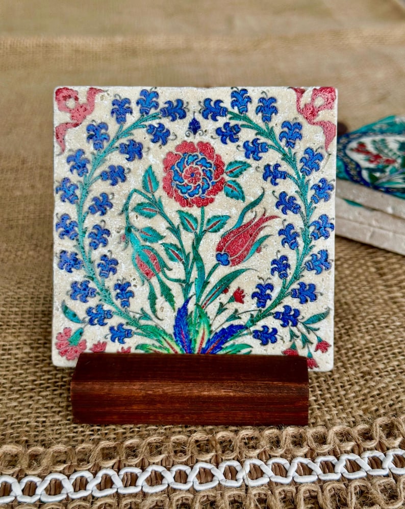 May include: Square ceramic coaster with a floral design in red, blue, and green on a cream background. The coaster is displayed on a dark brown wooden stand. Additional coasters are visible in the background.