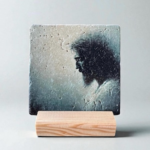 May include: A square, stone-like tile featuring a profile portrait of a man with long hair and a beard, set against a muted, textured background. The tile rests on a small, natural wood stand, creating a display piece.