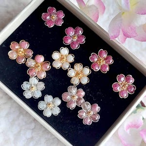 Puede incluir: Una colección de pendientes con forma de flor en una caja. Los pendientes presentan varios colores, incluyendo rosa, blanco y transparente, con detalles dorados. Están dispuestos sobre una superficie negra.