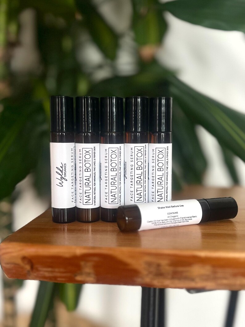 May include: Six small, black and white labeled bottles of face targeting serum called "Natural Botox" on a wooden surface. The bottles are arranged in a row, with the labels facing the viewer. The text on the labels reads "Face Targeting Serum Natural Botox A Natural Retinol for Wrinkles Fine Lines & Firming Skin".