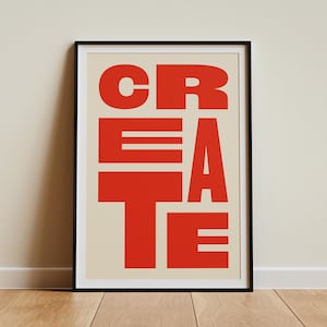 May include: A framed art print with the word "CREATE" in bold, red, block letters. The letters are arranged vertically and overlap, set against a cream background. The artwork is in a black frame and leans against a beige wall.