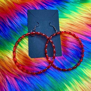 May include: A pair of red hoop earrings with a twisted design. The earrings are displayed on a black card against a vibrant, multicolored background of rainbow hues. The earrings have silver-colored hooks.