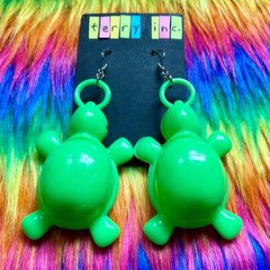 May include: A pair of bright lime green plastic turtle earrings attached to a black display card with the text terry inc. printed in colourful blocks. The earrings feature a rounded turtle shell design with four legs and a head, suspended from silver-toned metal hooks.