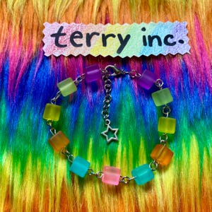 May include: A bracelet featuring translucent, multi-colored cube beads in shades of green, orange, pink, purple, and blue, connected by silver-toned metal links. The bracelet includes a silver-toned star charm and a tag reading terry inc.