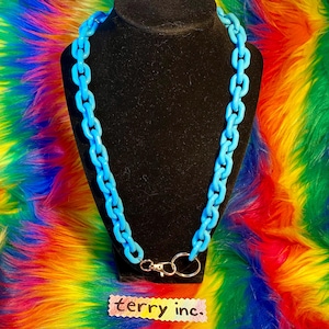 May include: A light blue chain necklace displayed on a black mannequin. The necklace has large, chunky links and a silver-colored clasp. The background is a vibrant, multicolored faux fur. A small tag at the bottom reads "terry inc."