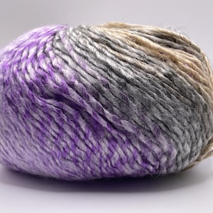 May include: A large ball of yarn with a gradient of colors, transitioning from purple to gray and tan. The yarn appears thick and textured, suitable for knitting or crocheting projects. The ball is sitting on a white surface.