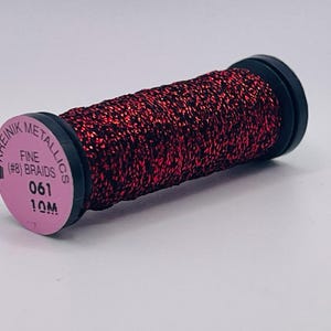 May include: A spool of Kreinik Fine (#8) Braids metallic thread in a deep red color. The spool features a pink label on the end with the text Kreinik Metallics, Fine (#8) Braids, 061, and 10M. The thread has a shimmering, textured appearance.