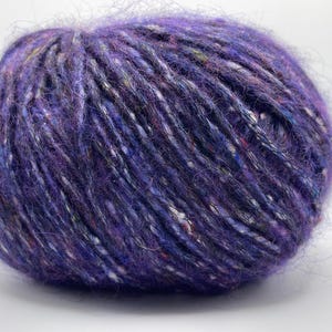 Peut inclure: Une pelote de laine violette avec des mouchetures blanches, bleues et d'autres couleurs. La laine a une texture douce et duveteuse, adaptée aux projets de tricot ou de crochet. La laine est d'une nuance de violet profond.