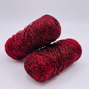 May include: Two spools of red yarn with a metallic sheen, ideal for knitting or crochet projects. The yarn has a deep red color with darker red and black accents, creating a textured appearance. The spools are positioned on a white background.