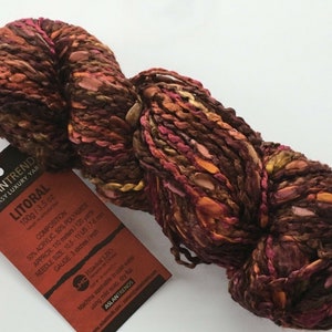 May include: A skein of Litoral yarn in a mix of brown, orange, pink, and gold colors. The yarn is made of 50% acrylic and 50% polyamide. The label says "100g / 3.5 oz" and "Litoral".