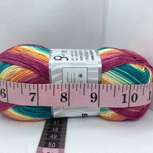 May include: A ball of yarn with a multi-coloured striped pattern in shades of pink, orange, yellow, and teal. The yarn is wrapped around a label with the text "GSC TH" and "info@gsc.th". The yarn is measured with a pink tape measure, showing a measurement of 20 cm.