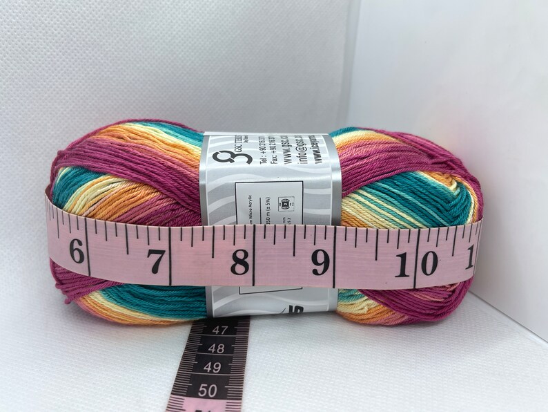 May include: A ball of yarn with a multi-coloured striped pattern in shades of pink, orange, yellow, and teal. The yarn is wrapped around a label with the text "GSC TH" and "info@gsc.th". The yarn is measured with a pink tape measure, showing a measurement of 20 cm.