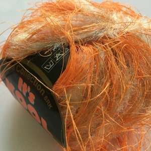 May include: A ball of orange and cream-colored yarn with long, textured strands. The yarn is wrapped around a label that reads "It's Wool" in orange text. The yarn appears to be soft and fluffy, suitable for knitting or crochet projects.