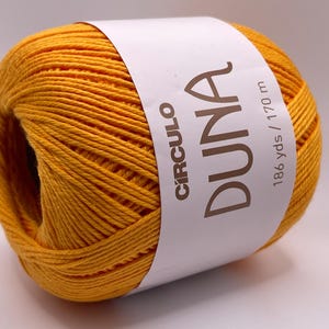 May include: A ball of yellow yarn with a white label wrapped around it. The label reads "CIRCULO DUNA" and "186 yds / 170 m". The yarn is a solid, bright yellow color, and the texture of the yarn is visible.