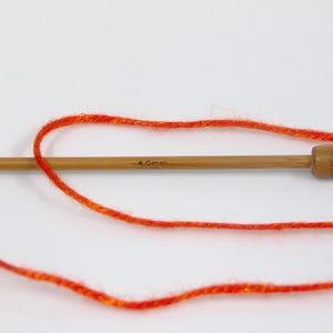 Puede incluir: Primer plano de una aguja de tejer de madera con el texto "4.0mm" y lana naranja brillante. La lana est&aacute; enrollada alrededor de la aguja, lo que sugiere un proyecto de manualidades. La aguja es de madera de color marr&oacute;n claro.