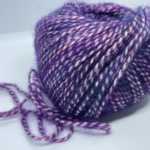May include: A skein of purple yarn with white and pink stripes and a silver shimmer. The yarn is soft and fluffy, and it would be perfect for knitting or crocheting.