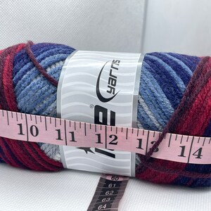 May include: A skein of blue, red, and white yarn with a label that reads "Big Yarns". The yarn is measured with a pink tape measure that reads 9 to 15 inches.