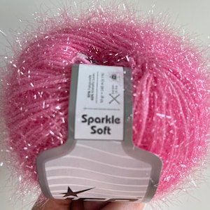 Può includere: Un gomitolo di filato Sparkle Soft rosa con un'etichetta bianca che dice "Sparkle Soft" e ha un logo a forma di stella. Il filato ha una consistenza soffice e un leggero luccichio.
