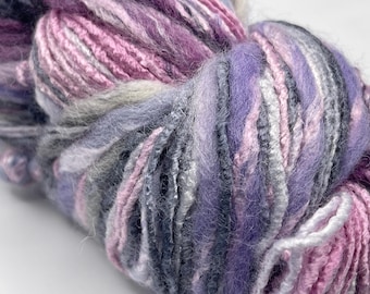 May include: A skein of yarn in shades of pink, purple, gray, and white. The yarn is soft and fluffy, and it has a subtle sheen.