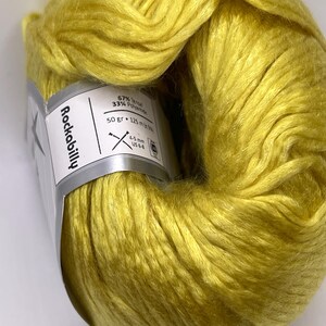 May include: A skein of bright yellow yarn with a sheen, wrapped in a white paper label that reads Rockabilly. The label specifies 67% Tencel and 33% Polyamide, a weight of 50 grams, a length of 125 meters, and a recommended needle size of 4-5 mm or US 6-8.