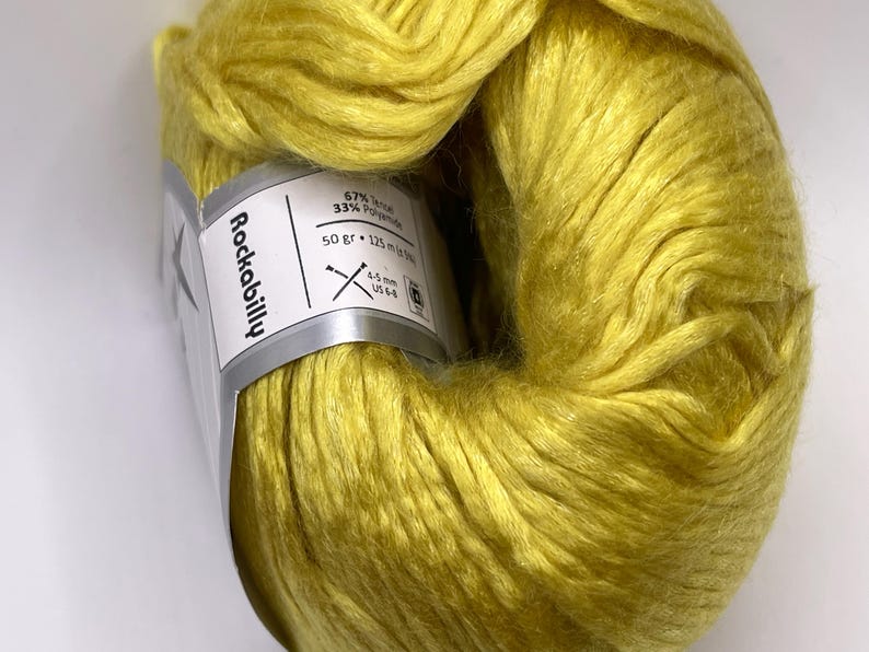 May include: A skein of bright yellow yarn with a sheen, wrapped in a white paper label that reads Rockabilly. The label specifies 67% Tencel and 33% Polyamide, a weight of 50 grams, a length of 125 meters, and a recommended needle size of 4-5 mm or US 6-8.