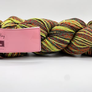 Puede incluir: Ovillo de lana Louisa Harding Marmo en una mezcla de colores que incluye verde, naranja y marrón. La lana está etiquetada con una etiqueta rosa que dice "Marmo" y "Marbled Piles of Colour".