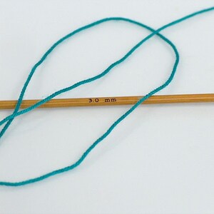 May include: Teal yarn wrapped around a 3.0 mm knitting needle.