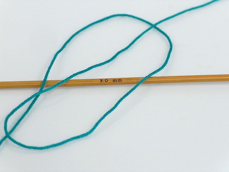 May include: Teal yarn wrapped around a 3.0 mm knitting needle.
